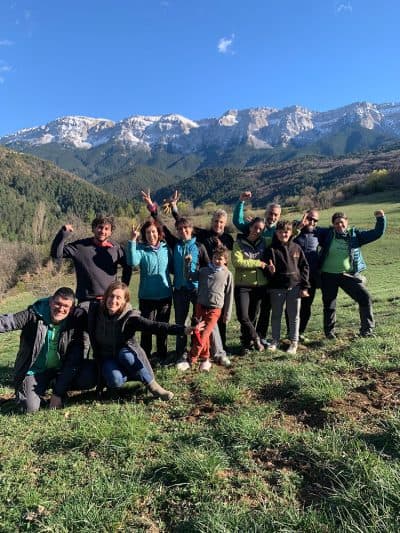 Grup de persones, incloent adults i nens, gaudint d’una jornada a la muntanya. Tots estan somrient i fent gestos alegres, amb els braços aixecats i signes de victòria.