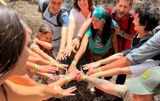 Persones de diferents edats reunides en un entorn natural, formant un cercle i unint les seves mans al centre en senyal d'unitat i col·laboració. Totes porten polseres verdes amb el color de la Fundació Miranda.