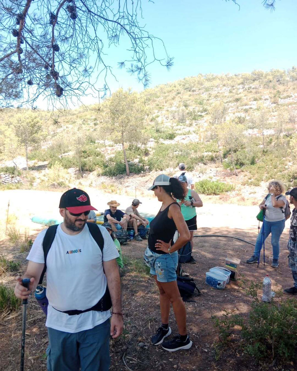 Una fotografia d'un grup de persones en un camí de terra sota l'ombra d'un arbre, amb vegetació i un turó al fons, algunes amb pals de senderisme i equipament d'exterior.