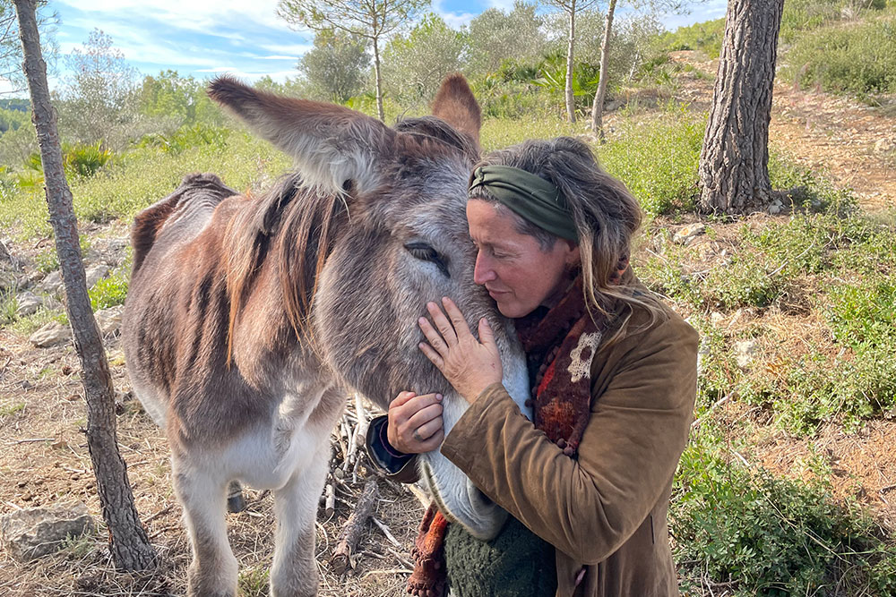 Una dona amb jaqueta color camell acarona i abraça el cap d'un ase bru-grisenc en un bosc assolellat. Tots dos tenen els ulls tancats, transmetent un profund moment de calma i connexió mútua.