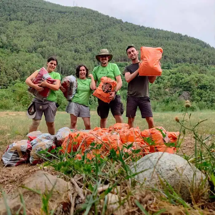 Un grup de quatre persones voluntàries, somriuen mentre sostenen grans sacs de color taronja i granat en un camp obert. Tres dels membres porten samarretes de color verd de la Fundació Miranda.