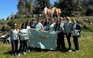 Grup nombrós de persones amb samarretes de color verd clar somrient i sostenint una bandera i bosses amb el logotip de Mandarin Oriental en un prat, amb diversos cavalls pasturant al turó que hi ha just darrere seu sota un cel blau.