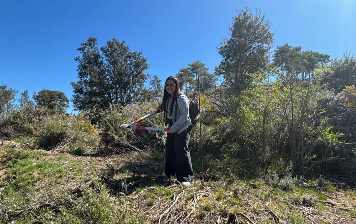 Team building-Mandarin Oriental-Fundació Miranda Dona somrient mentre utilitza unes tisores de podar de mango llarg per tallar arbustos en una zona de bosc baix, durant una jornada de voluntariat sota un cel blau clar.