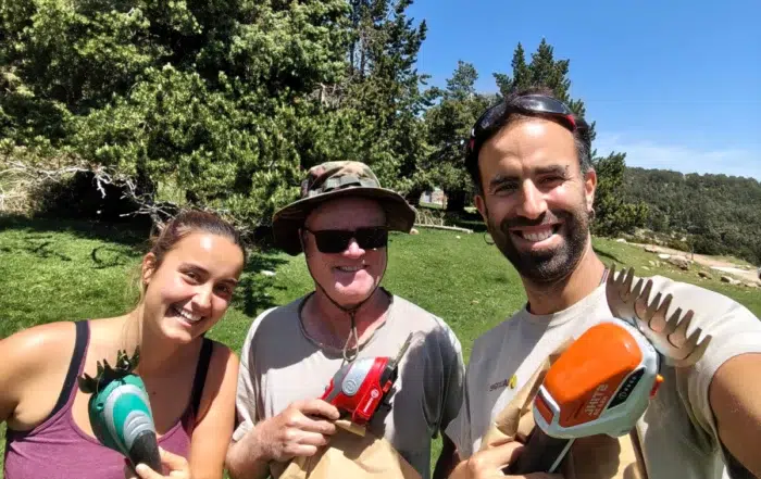 Tres persones somrients, equipades amb tisores elèctriques de jardineria i bosses de paper, posen per a una selfie en un entorn natural assolellat durant un treball de camp.