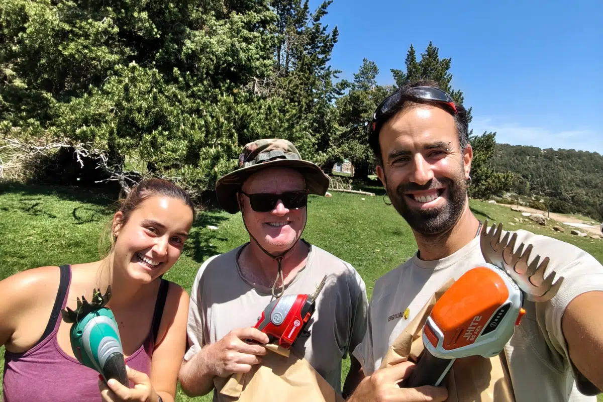 Recerca-Projecte GISKA Tres persones somrients, equipades amb tisores elèctriques de jardineria i bosses de paper, posen per a una selfie en un entorn natural assolellat durant un treball de camp.