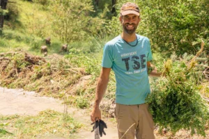 Un voluntari amb gorra i samarreta blava somriu a la càmera mentre subjecta branques i una eina al costat del curs d'aigua a l'espai natural de Collegats-Boumort.